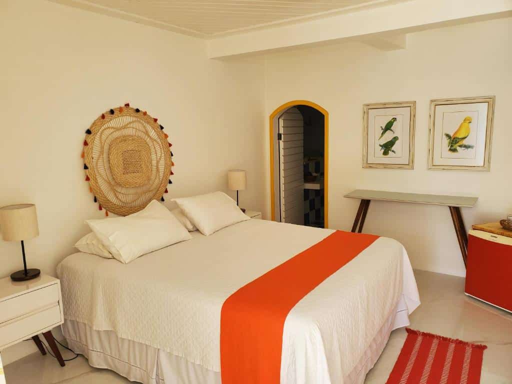 Quarto na Pousada Mestre Augusto com cama branca, colcha laranja, decoração de parede em tecido, duas gravuras de pássaros, escrivaninha e móveis neutros, para representar hotéis em Angra dos Reis.