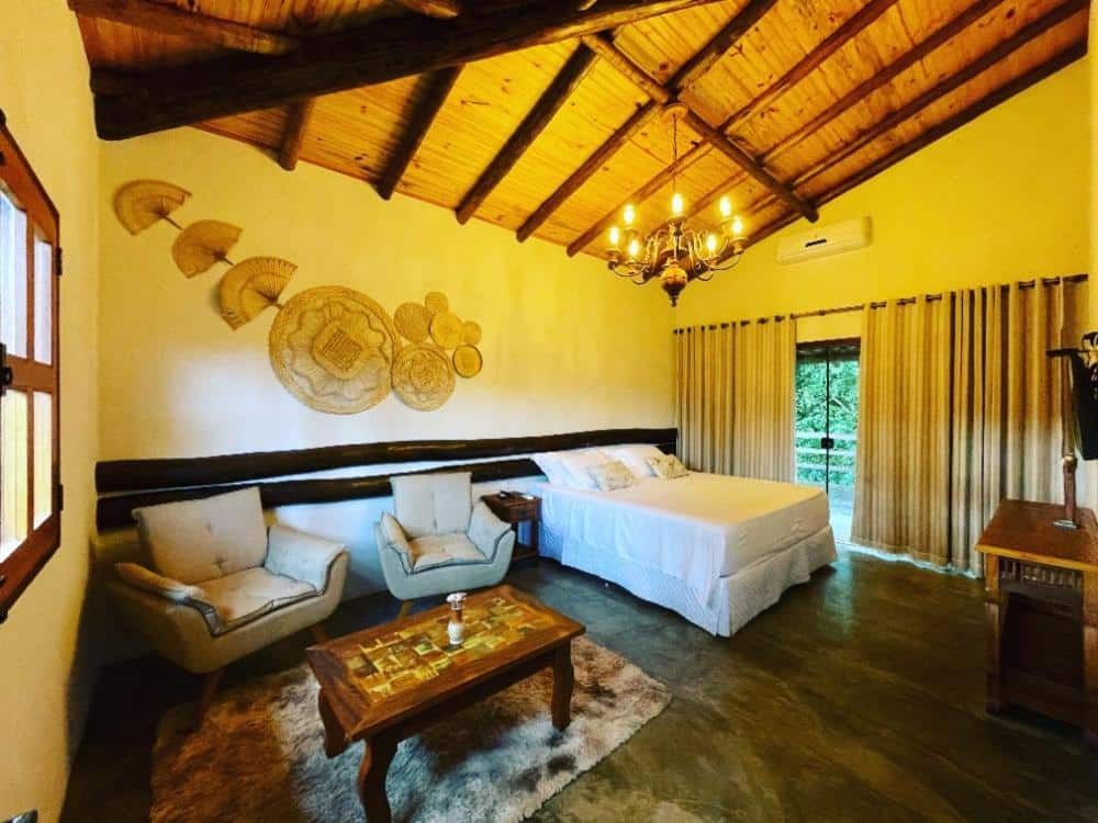 Um quarto rústico na Pousada Vale dos Diamantes com teto de madeira, cama de casal, duas poltronas, um tapete e um lustre, com cortinas cobrindo as janelas.