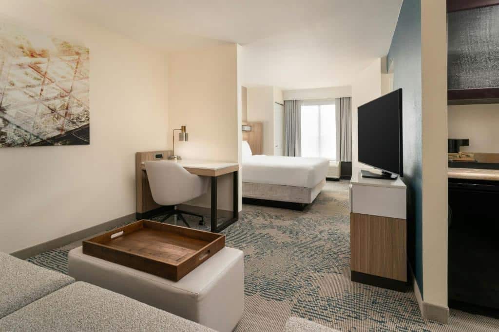Quarto moderno no SpringHill Suites by Marriott Tampa Westshore com cama, escrivaninha e cadeira, TV, sofá, pufe com bandeja e janela grande com luz natural, para representar onde ficar em Tampa.