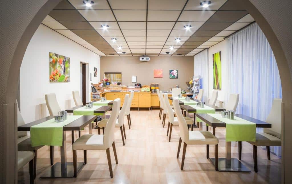 Uma sala de jantar moderna com oito mesas, cadeiras brancas, toalhas de mesa verdes e um bufê nos fundos, no Hotel-Pension Wild.