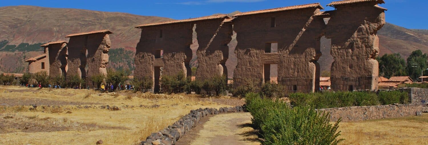 Ruínas de pedra com paredes altas e portas retangulares estão em uma paisagem seca e gramada com montanhas ao fundo na Rota do Sol saindo de Cusco até Puno
