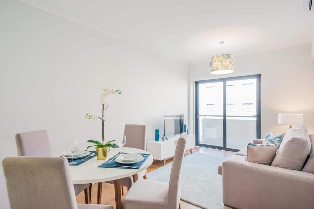 Área de estar e jantar bem iluminada com uma mesa redonda, quatro cadeiras, um sofá, TV e uma grande janela para uma varanda, no 137 LuxResidence.