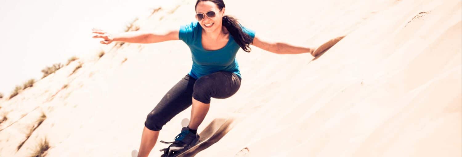 Uma mulher usando óculos escuros pratica sandboard em uma encosta arenosa, equilibrando-se com os braços estendidos e sorrindo no Tour de 4x4 pelo deserto de Chilca ou Marcahuasi