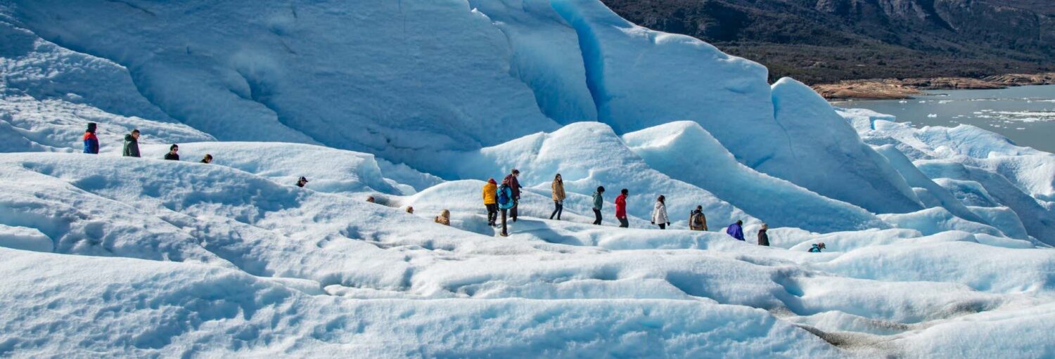 Um grupo de pessoas está ao ar livre perto de uma foto emoldurada, de frente para uma grande geleira em um corpo de água. Imagem para ilustrar post sobre o trekking pelo glaciar Perito Moreno.