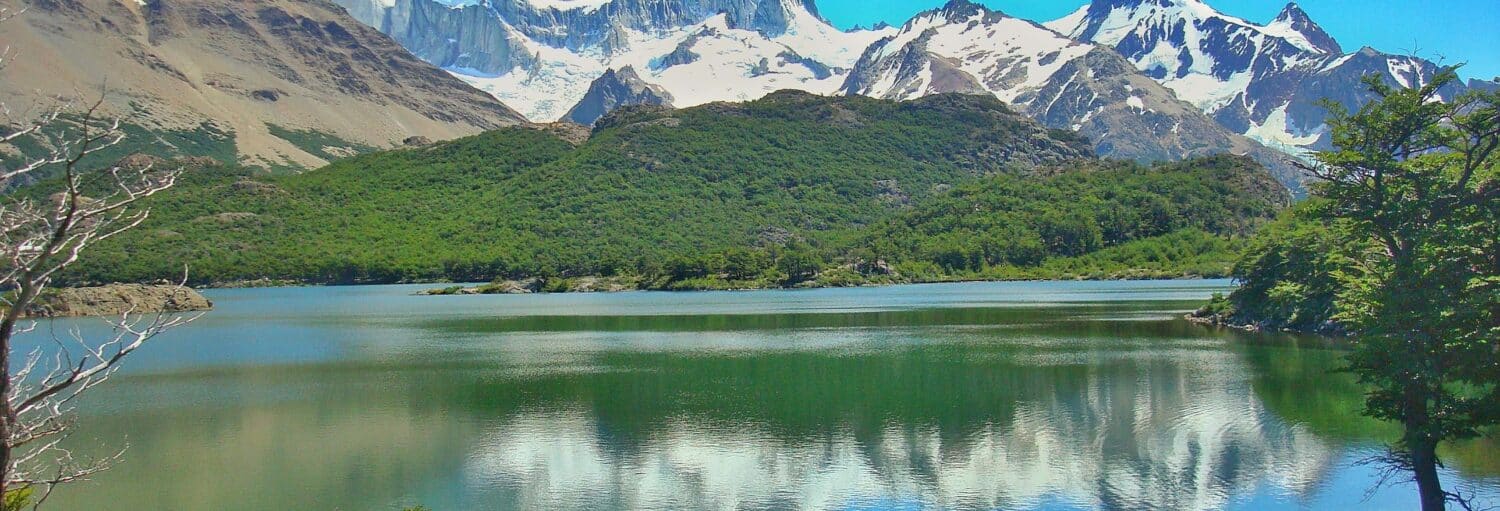 Montanhas cobertas de neve e colinas verdes refletidas em um lago calmo sob um céu azul claro, com algumas árvores em primeiro plano. Imagem para ilustrar post sobre a trilha pela Laguna Capri.