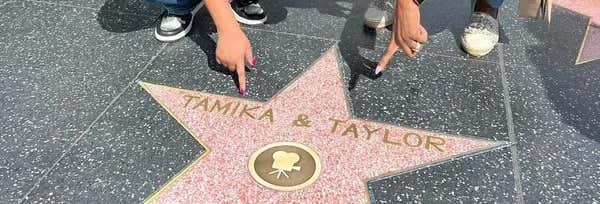 Duas pessoas apontam para uma estrela rosa da Calçada da Fama de Hollywood onde se lê "Tamika & Taylor" em uma calçada preta manchada. A foto é capa do post da Estrela na Calçada da Fama.