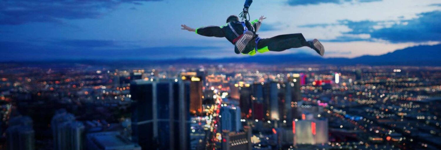 Uma pessoa em um arnês está pulando de bungee jump de um prédio alto ao anoitecer, com o horizonte da cidade ao fundo. Imagem para ilustrar post sobre SkyJump na The Strat Las Vegas.