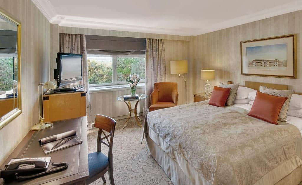 Quarto no InterContinental Wien com uma cama de casal, escrivaninha, cadeira, TV, poltrona, mesa lateral com flores e uma janela com cortinas, para representar onde ficar em Viena.