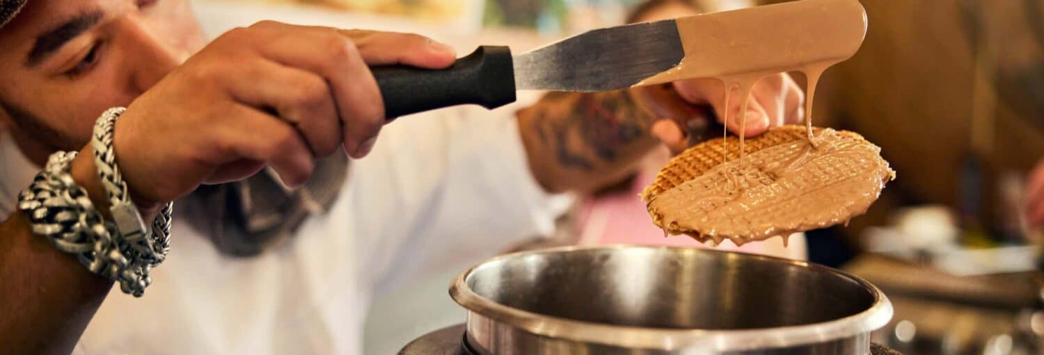Foco nas mãos de uma pessoa. A mão direita segura um waffle, enquanto a esquerda está com uma faca recheando-o com uma calda doce marrom. Oficina de stroopwafels em Amsterdam