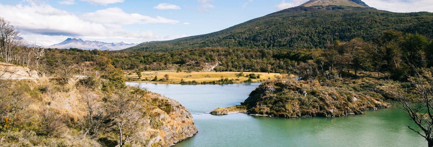 Um rio se curva em uma paisagem verde com colinas arborizadas e montanhas distantes sob um céu parcialmente nublado. Imagem para ilustrar post sobre o Parque Nacional Terra do Fogo + Trem do Fim do Mundo para cruzeiros.