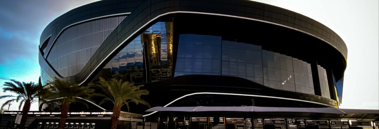 Um estádio moderno e preto com arquitetura curva, grandes painéis de vidro e palmeiras na frente. Imagem para ilustrar post sobre tour pelo Allegiant Stadium.