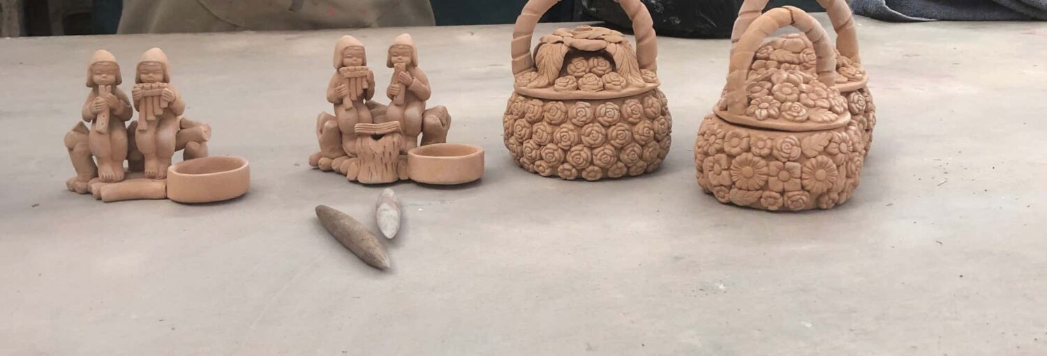 Três figuras de argila e duas cestas de argila com padrões florais são exibidas em uma mesa cinza com algumas ferramentas pequenas no Tour do artesanato ayacuchana