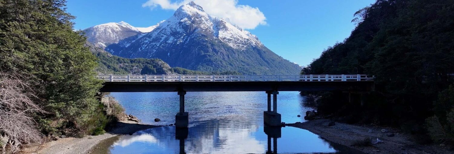 Uma ponte branca atravessa um lago calmo com uma montanha coberta de neve e colinas arborizadas ao fundo sob um céu azul no Tour de bicicleta pelo Circuito Chico