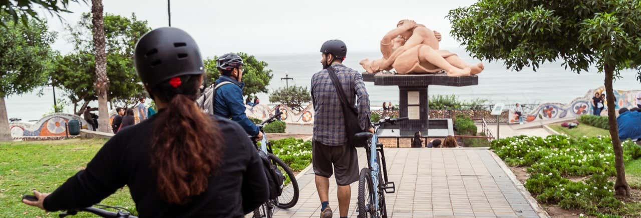 Várias pessoas com bicicletas olham para uma grande escultura de duas figuras se abraçando em um parque próximo ao oceano do Tour de bicicleta por San Isidro, Miraflores e Barranco