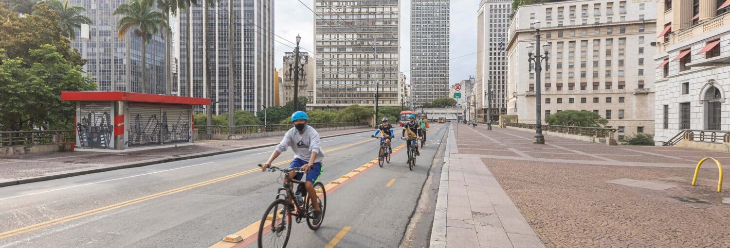 Quatro pessoas andam de bicicleta em uma ciclovia designada em uma área urbana com prédios altos e ruas vazias. A imagem ilustra o post sobre Tour de bicicleta por São Paulo.