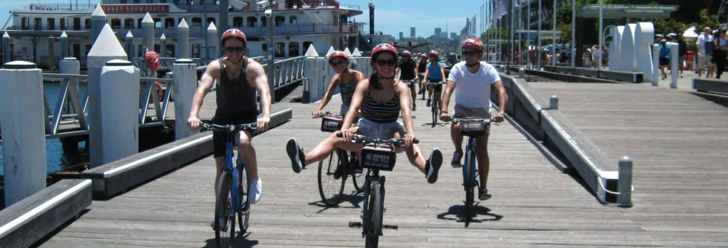 Quatro pessoas andam de bicicleta em uma doca de madeira ao lado de um barco fluvial e edifícios, com outras pessoas caminhando ao fundo. Imagem para ilustrar post sobre o tour de bicicleta por Sydney.