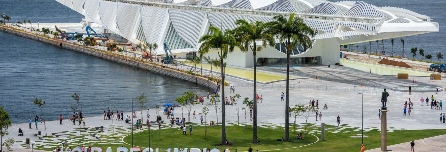 As pessoas caminham perto do Museum of Tomorrow, um moderno edifício branco à beira d'água com palmeiras e gramados verdes. a imagem ilustra o post sobre tour pelo Boulevard Olímpico + ingresso do AquaRio.