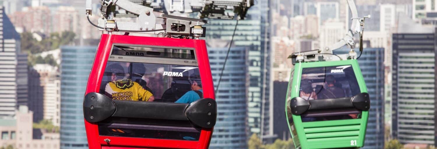 Dois teleféricos, um vermelho e um verde, viajam sobre fios acima de uma cidade com prédios altos ao fundo. A foto é capa do post do Tour pelo Cerro San Cristóbal.