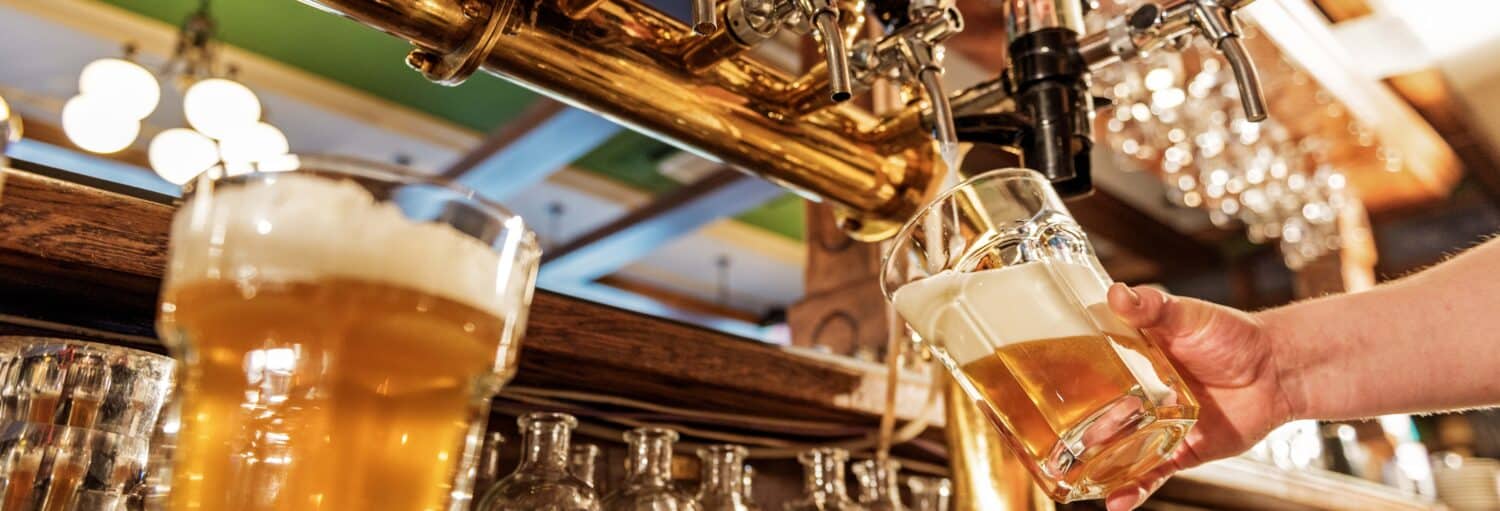 Uma mão despeja chope de uma torneira em um copo em um bar, com outro copo cheio e canecas ao fundo. Imagem para ilustrar post sobre tour da cerveja por Berlim.