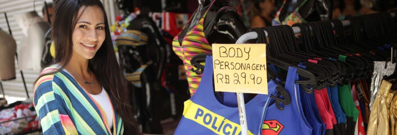 Mulher em pé ao lado de um cabideiro com roupas de banho e uma placa dizendo "Body Personagem R$ 29,90" em uma loja. A imagem ilustra o post sobre Tour de compras pelo Saara no RJ.
