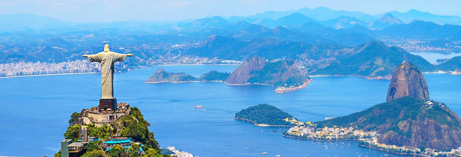 Vista aérea da estátua do Cristo Redentor com vista para o Rio de Janeiro, com montanhas e água ao fundo. A imagem ilustra o post sobre Cristo Redentor de trem + Tour pelo Rio.