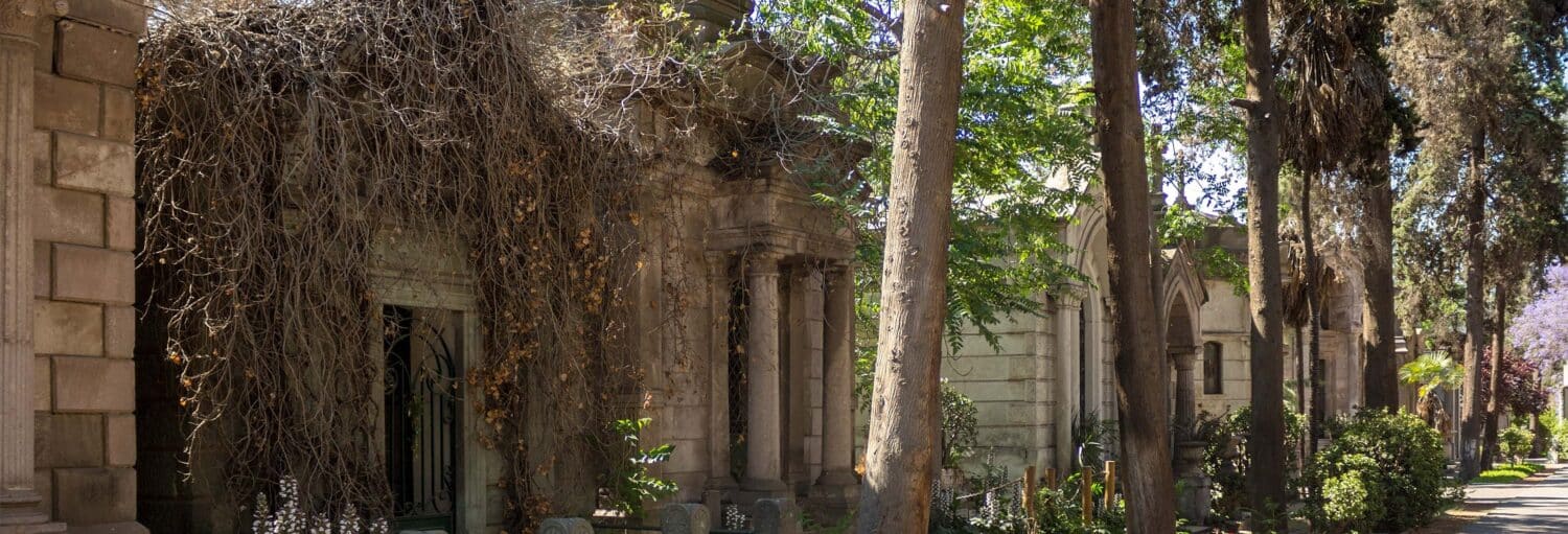 Fileira de mausoléus antigos de pedra com plantas e árvores crescidas ao longo de um caminho sombreado no cemitério. A foto é capa do post do Tour da ditadura de Pinochet.