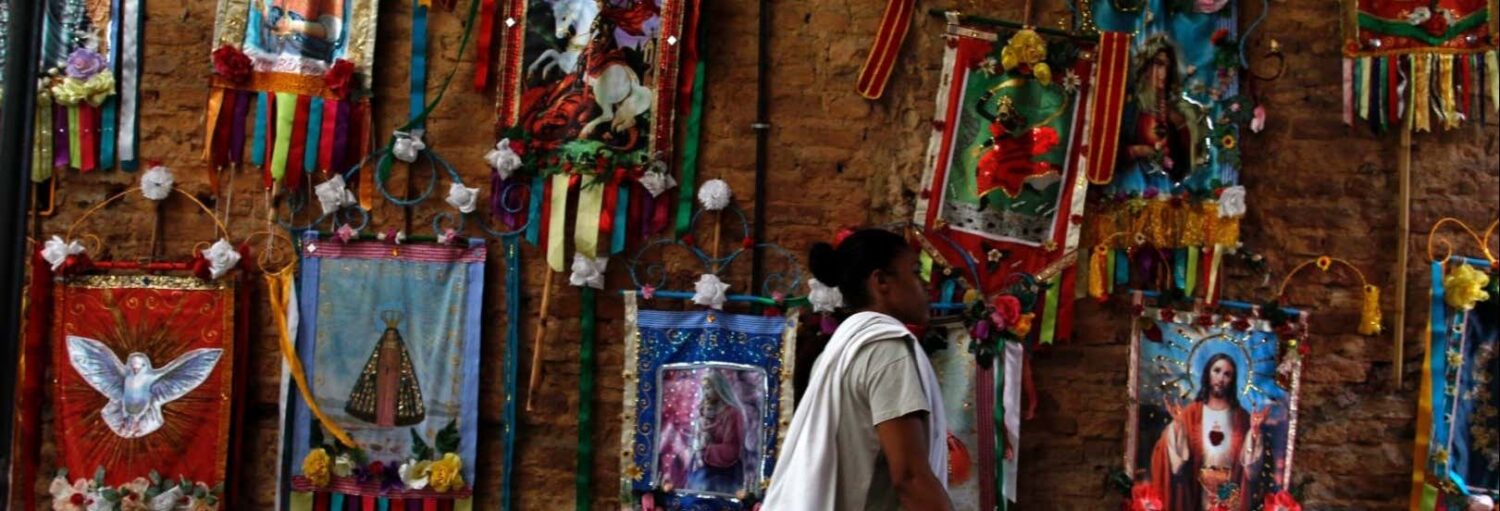 Uma pessoa está em frente a uma parede decorada com faixas e imagens religiosas coloridas. A imagem ilustra o post sobre Tour da diversidade cultural e religiosa em SP.