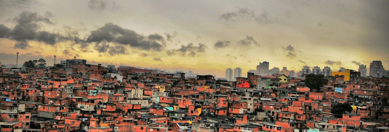 Uma favela densamente povoada com casas de tijolos vermelhos sob um céu nublado, com edifícios modernos da cidade visíveis ao fundo. A imagem ilustra o post sobre Tour privado pela favela de Paraisópolis.