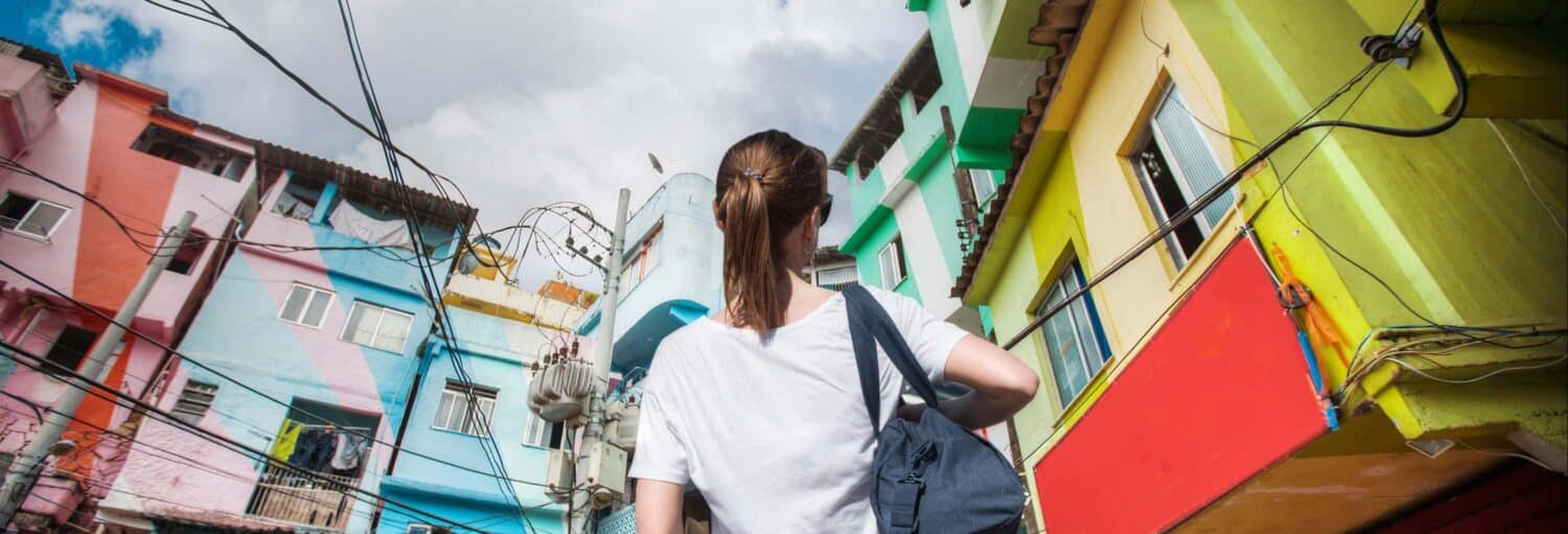 Uma pessoa com uma bolsa olha para edifícios coloridos e fios suspensos sob um céu parcialmente nublado. A imagem ilustra o post sobre tour pela favela Santa Marta.