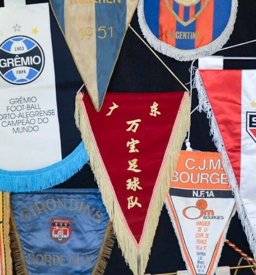 Vários galhardetes e bandeiras de clubes de futebol internacionais exibidos juntos em uma parede ou quadro. A imagem ilustra o post sobre Tour privado do futebol por São Paulo.