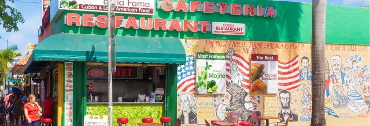 Vista da rua de um restaurante cubano com assentos ao ar livre, placas coloridas, murais e um toldo verde. A imagem ilustra o post sobre Tour gastronômico por Little Havana.