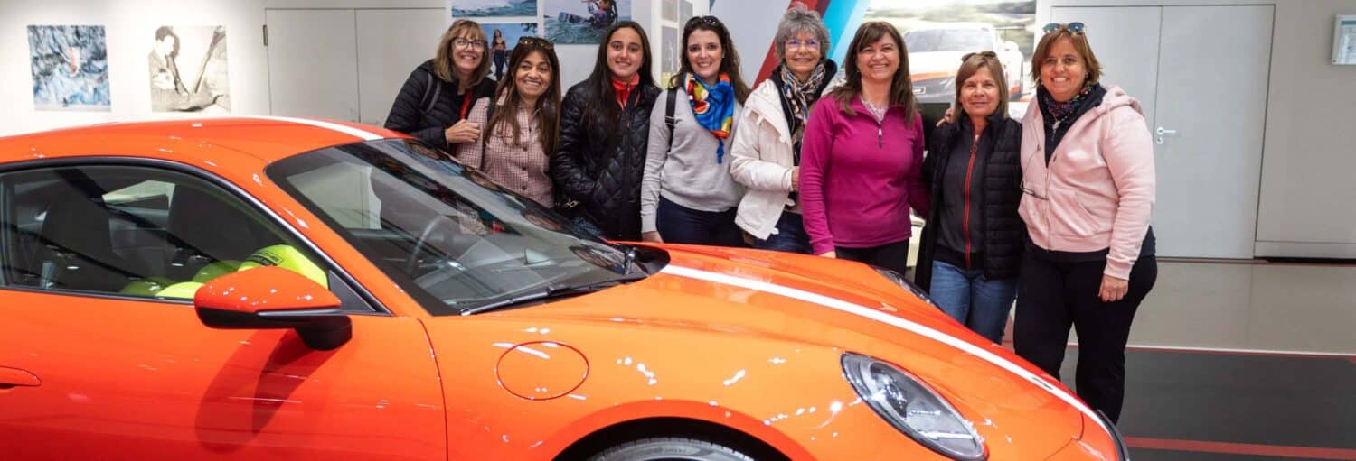 Nove mulheres estão juntas e sorriem atrás de um carro esportivo laranja em um ambiente interno bem iluminado com arte nas paredes. Imagem para ilustrar post sobre tour da história do automóvel por Berlim.