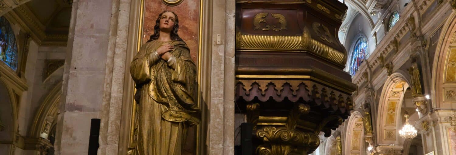 Uma estátua de uma mulher com as mãos dobradas em um interior de igreja ricamente decorado e ornamentado. A foto é capa do post do Tour pelas igrejas de Santiago.