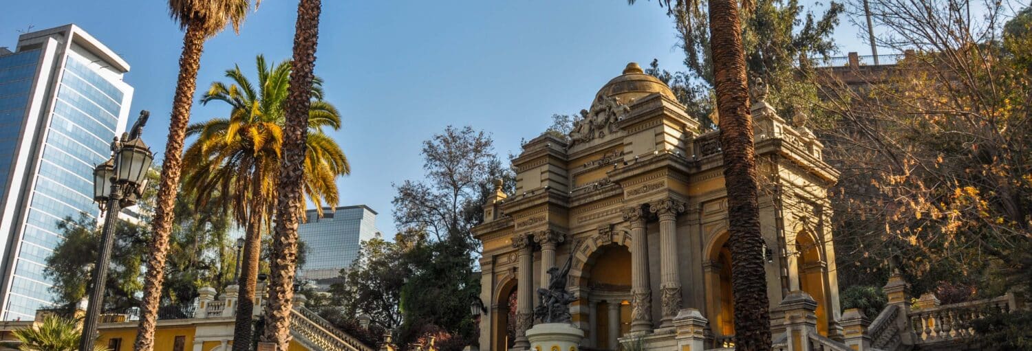 Edifício histórico de pedra com arcos e esculturas, cercado por palmeiras, com edifícios modernos ao fundo. A foto é capa do post do Tour por Lastarria e cerro Santa Lucía.