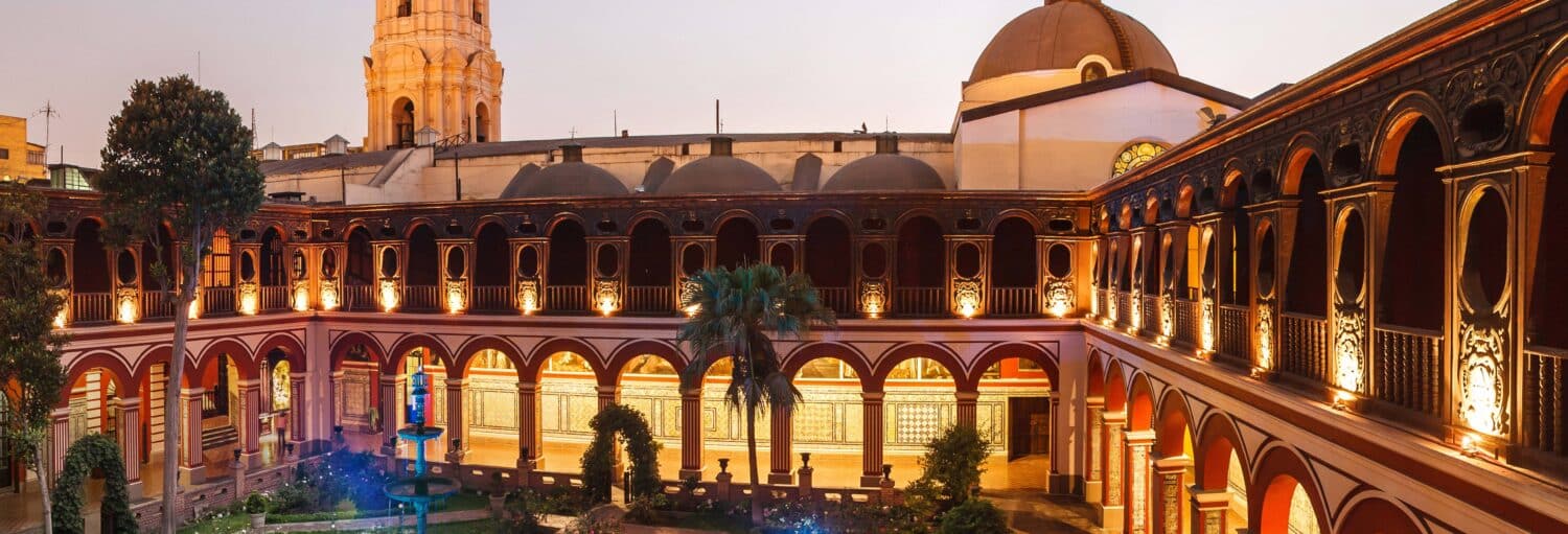 Pátio em estilo colonial com passarelas em arco, jardim central e um alto campanário ao fundo ao anoitecer no Tour pela Lima Colonial e pelo Bairro Chinês