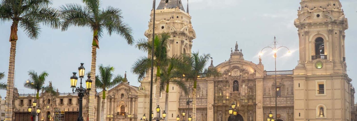 Catedral em estilo colonial com duas torres, palmeiras e lâmpadas de rua ornamentadas em uma praça da cidade ao anoitecer do Tour pela Lima colonial + Circuito Mágico da Água