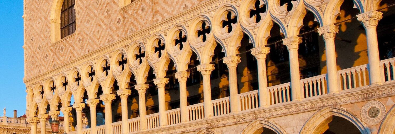 Fachada de pedra com janelas e colunas em arco, apresentando padrões geométricos e detalhes ornamentados sob luz solar quente. A imagem ilustra o post sobre Tour dos mistérios e lendas do Palácio Ducal.