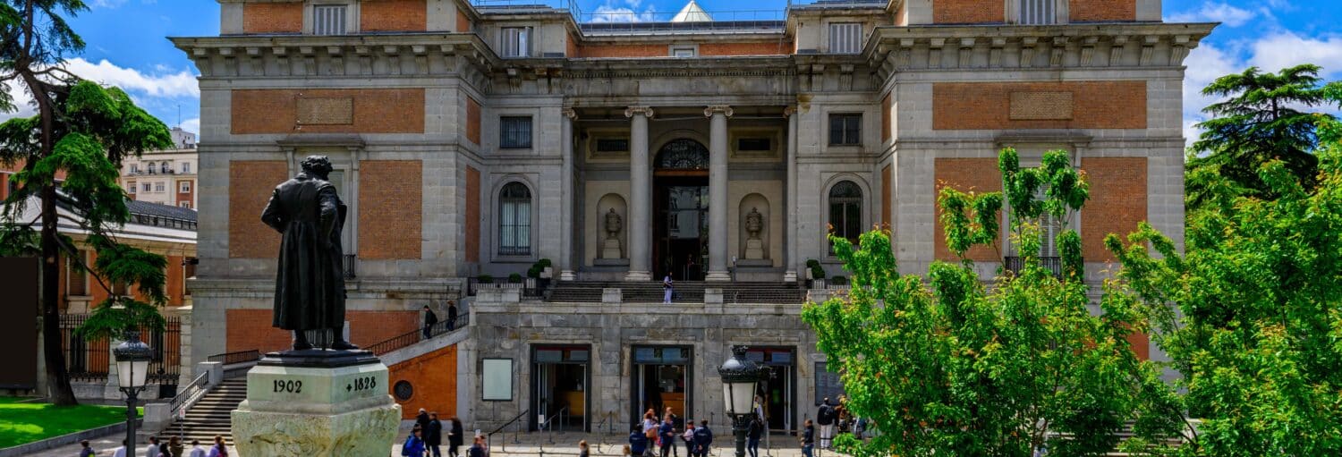 Um grande edifício de tijolos e pedras com colunas, uma estátua na frente e pessoas caminhando nos degraus e na praça. A imagem ilustra o post sobre Tour do Museu do Prado para crianças.