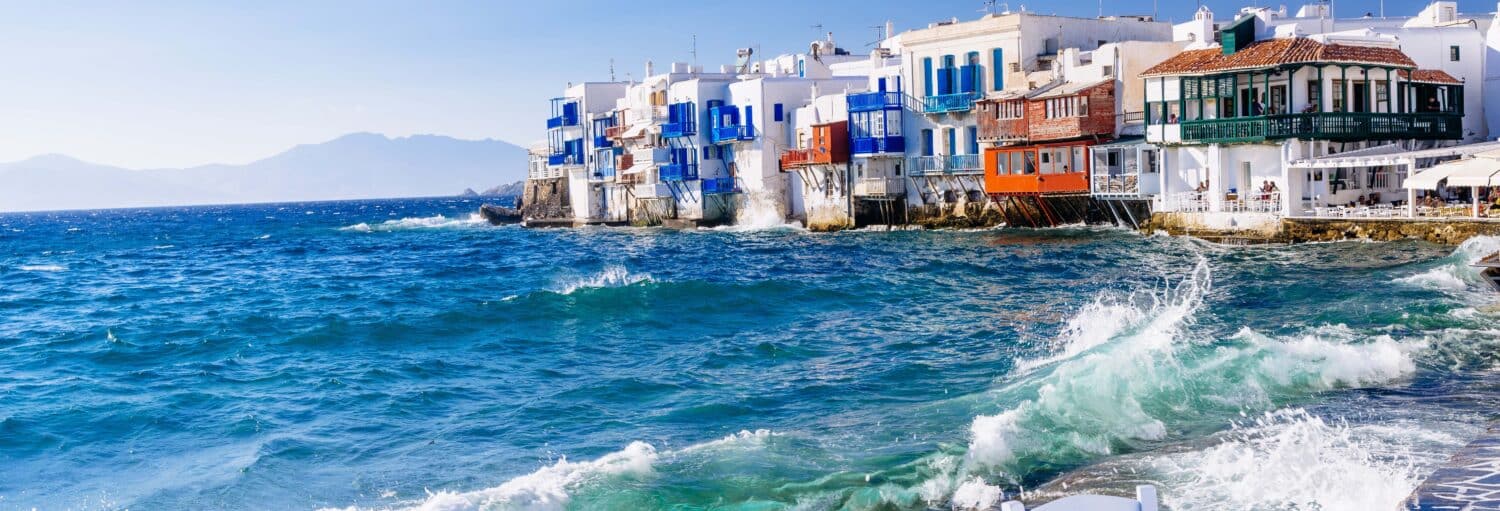 Casas coloridas à beira-mar com varandas se alinham à beira de um mar ondulado sob um céu azul claro, com montanhas ao fundo. A imagem ilustra o post sobre Tour de Mykonos para cruzeiros.