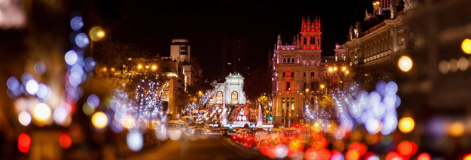 Uma rua da cidade à noite com luzes coloridas, tráfego embaçado e prédios históricos iluminados ao fundo. A imagem ilustra o post sobre Tour natalino de tuk tuk por Madrid.