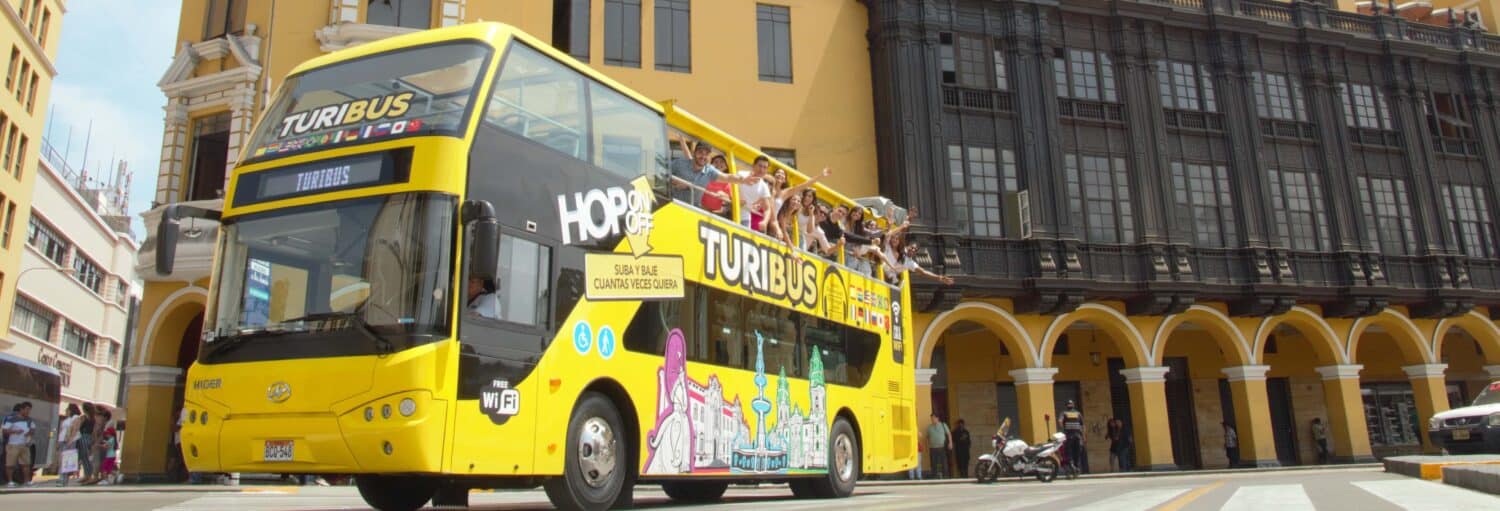 Um ônibus de turismo amarelo brilhante de dois andares passa por prédios históricos com pessoas no convés superior aberto no Tour panorâmico por Lima