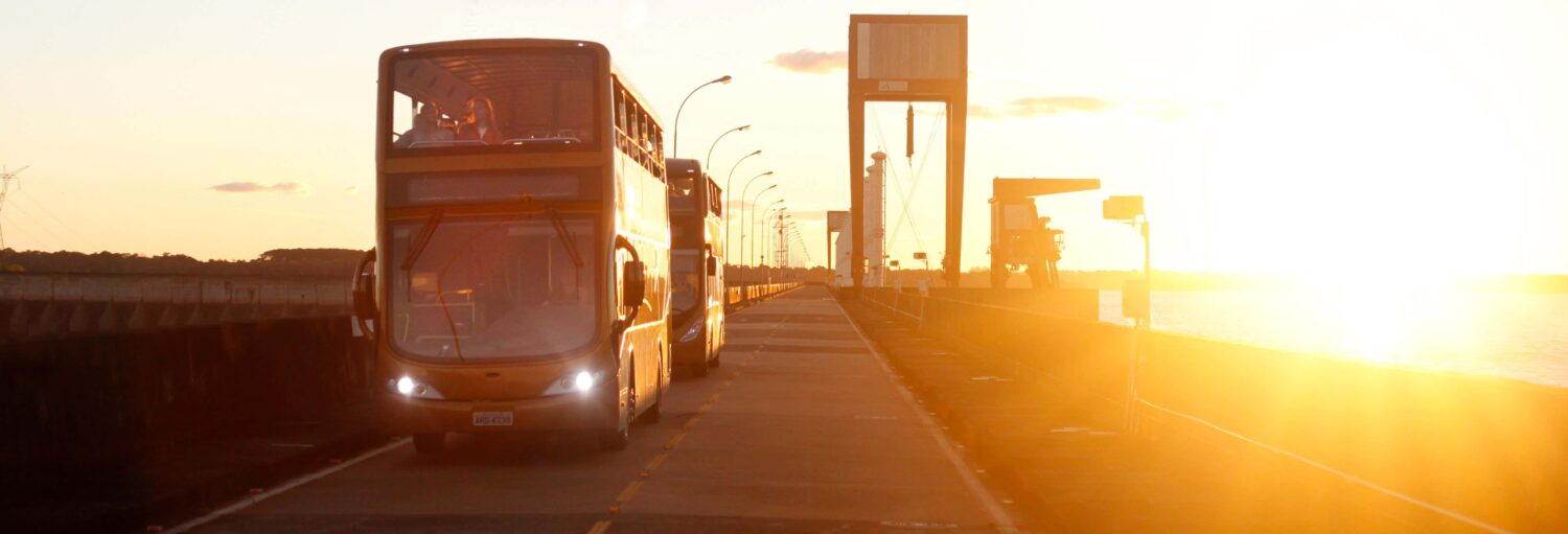 Dois ônibus de dois andares circulam por uma estrada ao lado da água ao pôr do sol, com a luz do sol brilhante ao fundo. - Tour panorâmico pela Usina de Itaipu