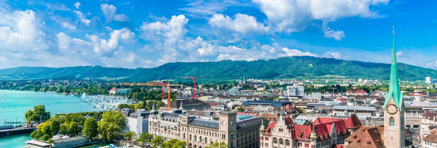 Uma vista panorâmica de Zurique com o lago, edifícios históricos e colinas verdes sob um céu azul com nuvens. Imagem para ilustrar post sobre o tour panorâmico por Zurique.