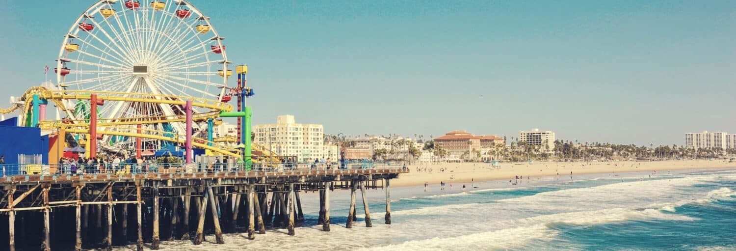 Uma grande roda-gigante e brinquedos de diversão em um píer têm vista para uma praia de areia com ondas e edifícios ao fundo. A foto é capa do post do Tour pelas melhores praias da Califórnia.