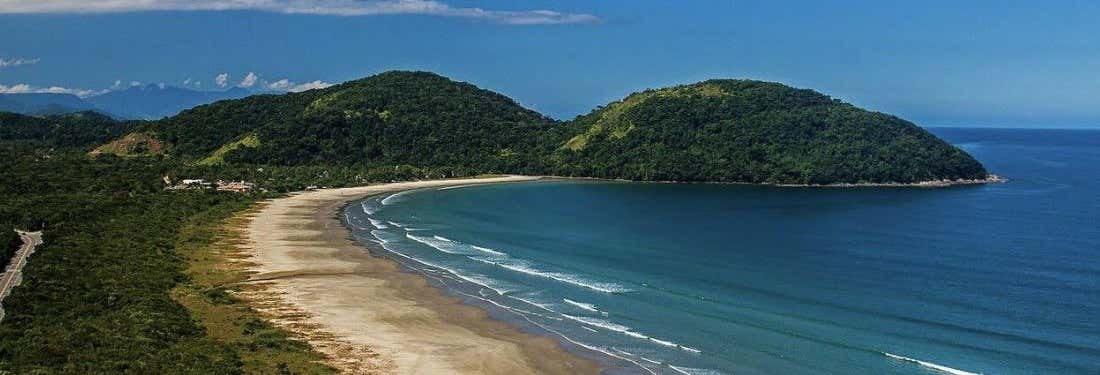 Uma ampla praia de areia se curva ao longo da costa, cercada por colinas verdes e um oceano azul calmo sob um céu claro. A imagem ilustra o post sobre tour pelas praias de São Sebastião saindo de SP.