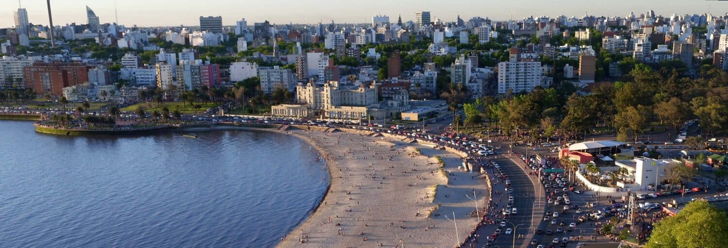 Vista aérea de uma praia urbana com pessoas na areia, árvores e vários edifícios ao fundo. Imagem para ilustrar post sobre tour privado por Montevidéu.