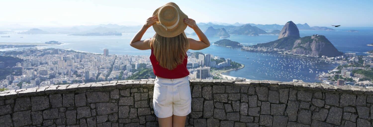 Pessoa de blusa vermelha e bermuda branca segurando um chapéu, com vista para uma cidade e uma baía com montanhas ao fundo. A imagem ilustra o post sobre Tour privado no Rio de Janeiro em português.