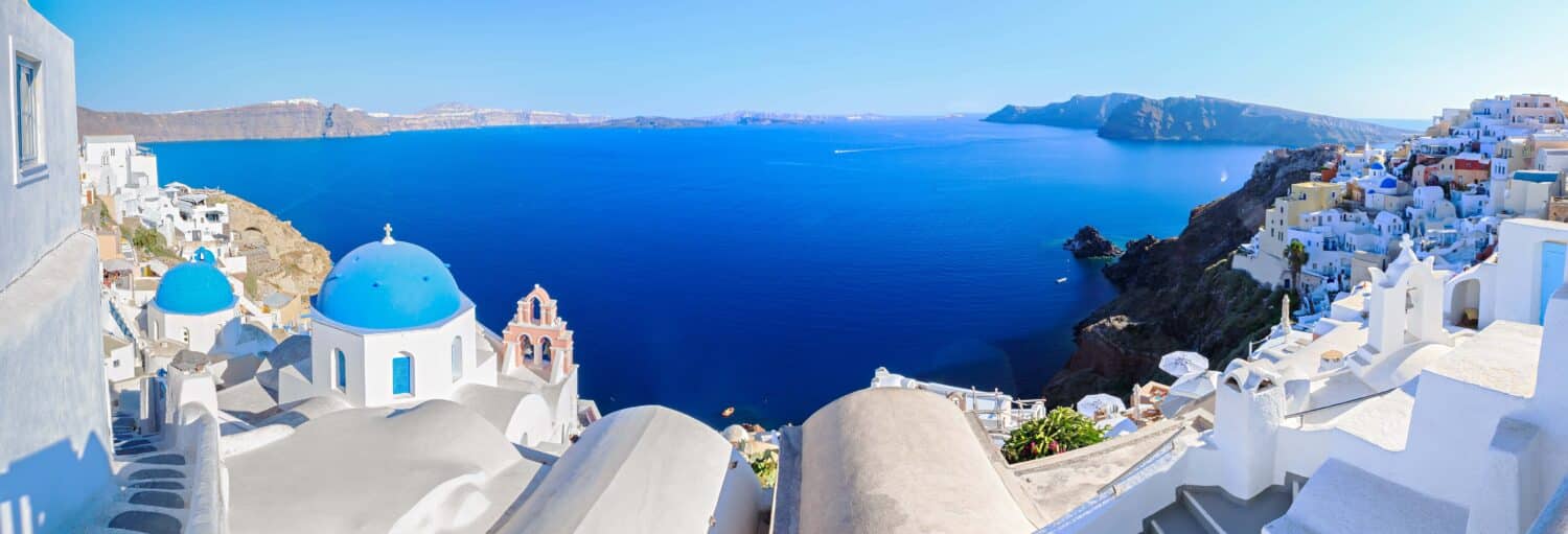 Vista panorâmica de Santorini com edifícios brancos, cúpulas azuis e o Mar Egeu sob um céu claro. A imagem ilustra o post sobre Tour privado por Santorini.