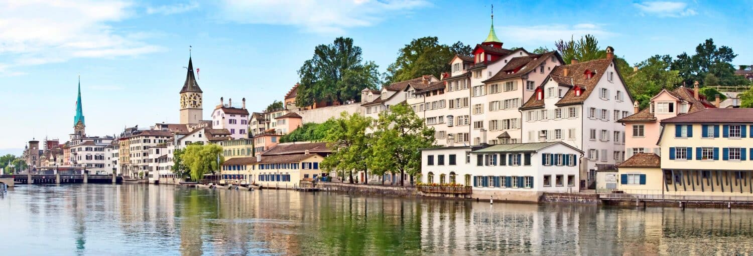 Fileira de edifícios históricos ao longo do rio Limmat, em Zurique, Suíça, sob um céu azul com alguma vegetação. Imagem para ilustrar post sobre tour privado por Zurique com guia em português.