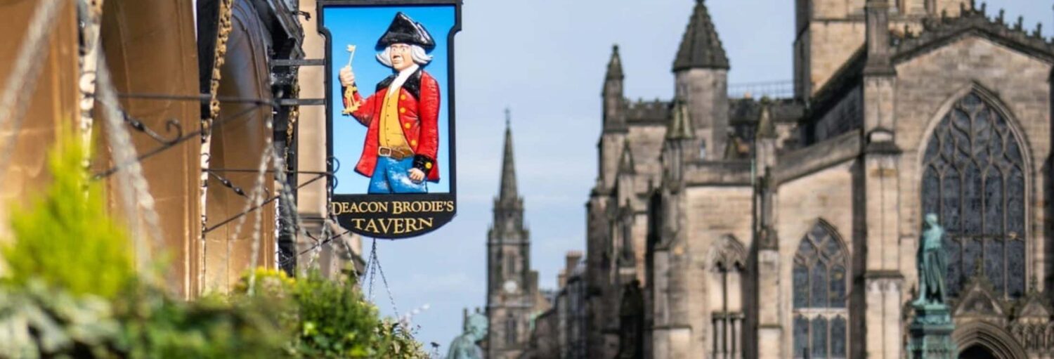 Uma placa suspensa da Deacon Brodie's Tavern com edifícios históricos de pedra e uma igreja ao fundo. A foto é capa do post do Tour pelos pubs históricos de Edimburgo.